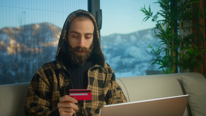 Bearded man in hoodie enters credit card number into laptop to online purchases. Man making easy...