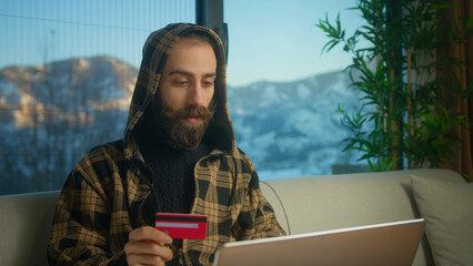 Young bearded man in hoodie enters credit card number into laptop to online purchases. Man making...
