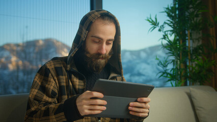 Bearded young man wearing hoodie playing video game on a tablet computer while sitting on sofa in...
