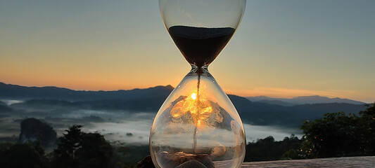 A hourglass (with falling sand) on a wooden wet table with sea of fog and mountain range silhouette...