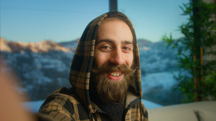 Bearded young man wearing hoodie making selfie or video call while sitting on the couch in living room. Male speaking on video call, using smartphone talking at camera webcam online