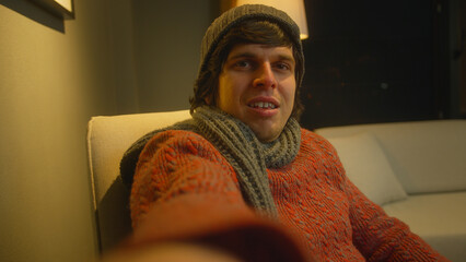 Young man wear scarf and beanie making having video call while sitting on armchair in living room. Man speaking on video call, using smartphone talking at camera online, selfie call