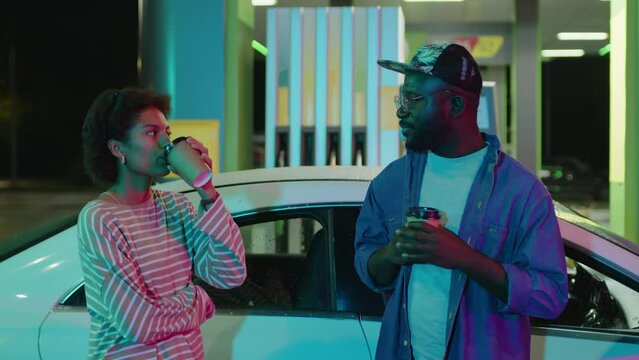 Medium footage of African American friends or fellow road trippers drinking coffee at neon lit petrol station and talking near white modern car at night