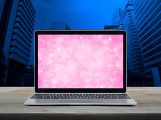 Modern laptop computer with pink love heart screen on wooden table over office city tower and...