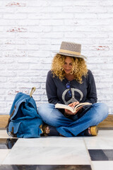 Alternative trendy travel people lifestyle beautiful curly fashion adult woman sit down on the floor and read a booko with her blue backpack nera her - white bricks background