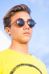 Handsome young boy man with blonde hair and black sunglasses stand and pose with yellow shirt and blue sky background - caucasian guy summer portrait