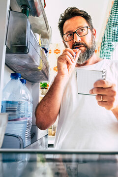 Adult caucasian man taking note list of food looking inside the open fridge at home - kitchen activity and alternative point of view - market shopping and quarantine lockdown stayhome coronavirus time