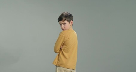 Offended little kid boy, sad stressed child looking at camera standing with his back isolated on grey background. Upset facial expression. Children problems concept
