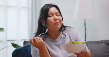 A chubby Asian woman enjoy eating salad while relaxing on the sofa at home.