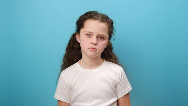 Portrait of unhappy preteen girl child looking at camera with sad upset expression, being in bad mood, wearing white casual t-shirt, posing isolated over plain blue color background wall in studio