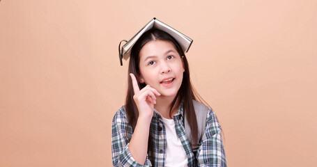 Cute school girl is carrying a school bag and holding a notebook on her head. Isolated on brown background in studio.