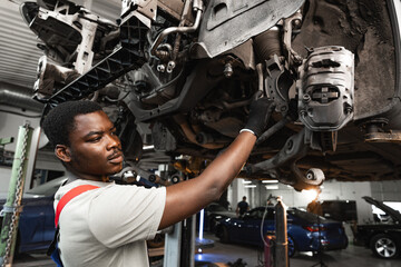 Young African mechanic in uniform working under the car in car service center