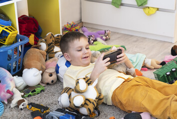 Child plays with phone among many scattered toys at home. Children's room is a mess. Dirty house. Children relaxing at home, video game addiction. Selective focus.