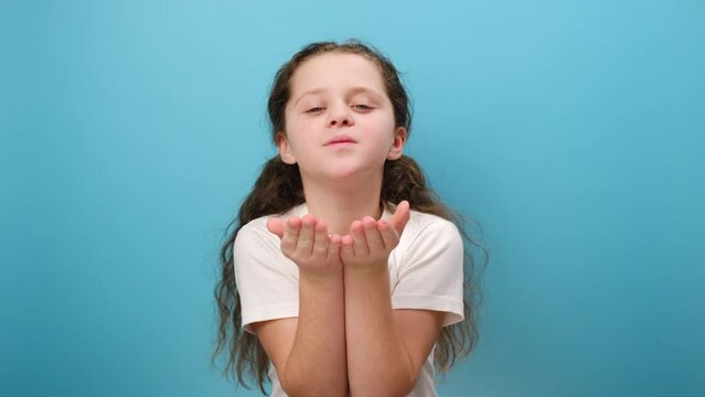 Portrait of charming cute little girl child sending air kisses to camera, keeping lips pout, looking at camera, wearing white casual t-shirt, posing isolated over blue color background wall in studio
