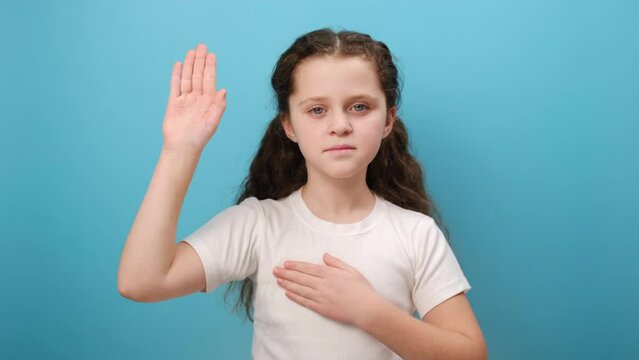 Portrait of serious preteen girl child raising her palm to take oath, child swearing to tell only truth, wearing white t-shirt, posing isolated over plain blue color background wall in studio