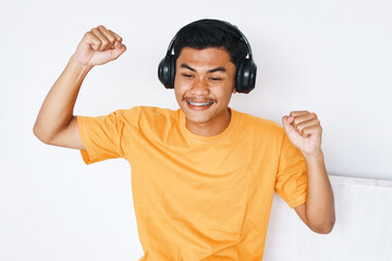 Fototapeta premium Happy Asian male student with braces teeth raising hands dancing while listening music from headphones