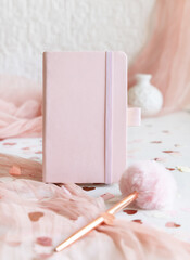 Pink hardcover notebook near hearts, pen, tulle and vase on white table close up, textbook mockup
