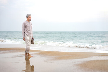 asian senior man barefoot,holding shoes walking on sand beach in the morning,healthy elderly male...