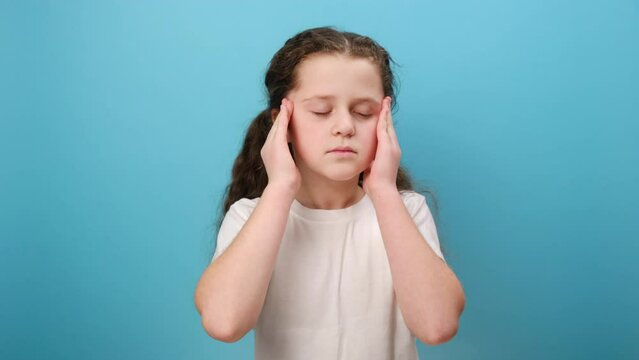 Portrait of unhappy little girl clutching temples feeling intense headache, child suffering migraine, medical problem, wearing white t-shirt, posing isolated on blue color background wall in studio