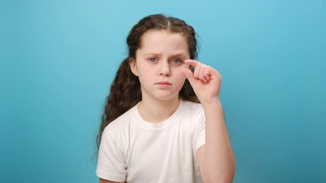 Portrait of little girl doing small gesture, looking with displeased imploring expression, shows minimum, wearing white casual style t-shirt, posing isolated over blue color background wall in studio