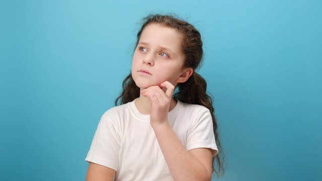 Portrait of adorable thoughtful preteen girl child stands holds chin, looking away with pensive expression, wearing casual white t-shirt, posing isolated over blue color background wall in studio