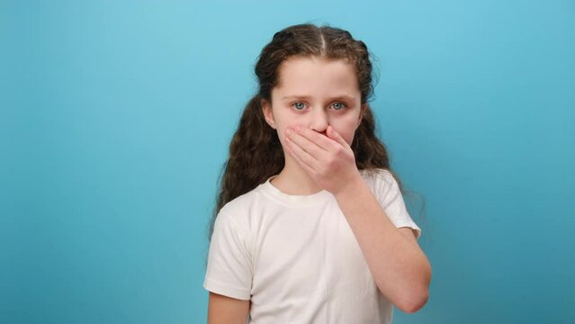 Portrait of worried little girl kid covering mouth with hand, afraid to say secret, child terrified to speak, wearing white t-shirt, posing isolated over plain blue color background wall in studio