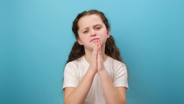 Portrait of little girl child looks up praying to God with pleading eyes, making childhood wish and hoping, wearing white casual t-shirt, posing isolated over blue color background wall in studio