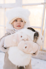 Closeup portrait of happy little child boy in warm clothes hugging white funny rabbit standing by farm house on sunny winter day, looking at camera. Concept of excursion to eco-farm, life in village.