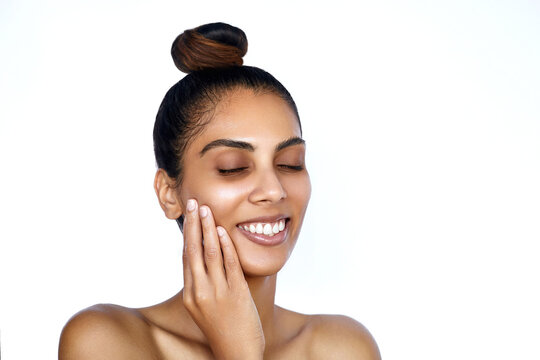  South asian young woman with closed eyes beauty skincare portrait. Beautiful indian model with a smooth bright hydrated skin and hair bun is posing with a chin look and smiling.
