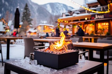 A cozy fire pit with a warm flame in a snowy outdoor setting at dusk, surrounded by festive lights and mountains. - obrazy, fototapety, plakaty
