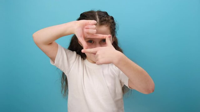 Portrait of preteen girl child looking through photo frame hand gesture, focusing picture, observing world, wearing white t-shirt, posing isolated over plain blue color background wall in studio