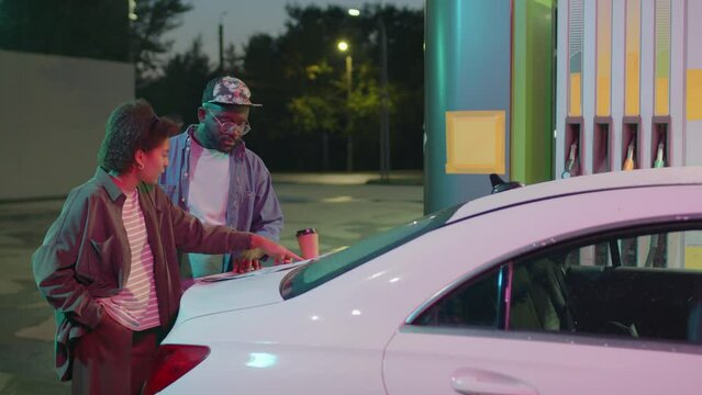 Side full medium shot of African American fellow road trippers deciding on route studying country map on car hood at petrol station at night