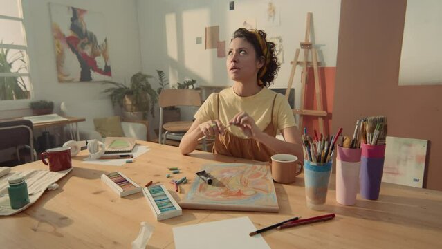 Medium shot of young biracial female art teacher sitting at table in workshop, with sketchbook, pastel, brushes and paints, and beginning painting lesson while filming social media content