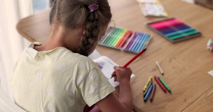 Concentrated girl draws picture with colored felt-tip pens sitting at wooden table in living room. Expressing creativity in childhood. Moments from childhood