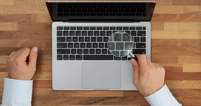Hand holds magnifying glass, looks through magnifying glass at keys of laptop top view