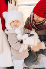 Vertical portrait of happy adorable son with loving family in warm clothes hugging white funny rabbit standing by house on sunny winter day. Concept of excursion to eco-farm, life in village.