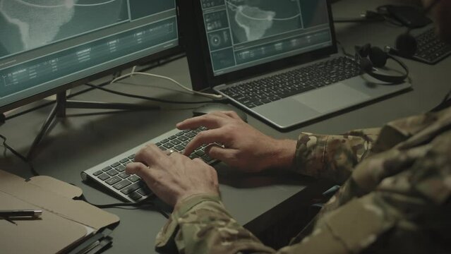 Over shoulder of military officer in camouflage uniform looking at satellite map on computer and laptop displays, typing and sending report via headset with mic while working in office at night