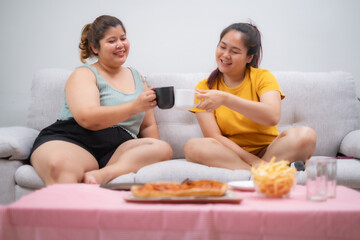 Obraz na płótnie Canvas Two Young Asian beautiful women drinking a cup of coffee with blur Pizza and french fries, unhealthy lifestyle, eating junk meal. overweight, obesity.