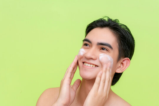 A young asian man feels happy while gently massaging exfoliating facial scrub cream to his face using his fingers. Skin care and hygiene concepts. Isolated on a light green backdrop.