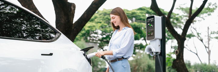 Young woman recharge EV electric vehicle's battery from EV charging station in outdoor green city...