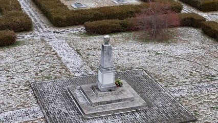 Overhead shot of a snowy WWII cemetery with a central obelisk, gravestones spread around, sun peeking through clouds.