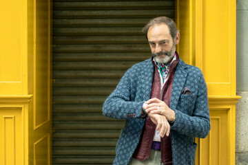 Adult man with beard leaning on a yellow wall waiting while looking at the time on his watch