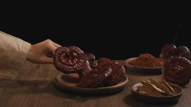 A hand is holding a lingzhi mushroom. Ganoderma mushrooms, sliced lingzhi mushrooms and lingzhi mushroom powder are displayed on wooden plates on a wooden table with a black background.