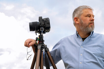Photographer at work, gray hair and full beard, 56 years old, light shirt, glasses set off in hand, 35mm SLR camera on tripod, gray overcast sky - Powered by Adobe