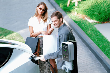 Young couple travel with EV electric car charging in green sustainable city outdoor garden in...