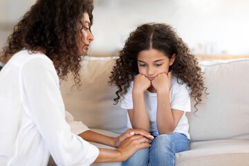 Worried mother calming unhappy preteen daughter, talking and helping with problems, sitting on sofa in living room interior. Family support and mother care