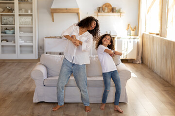Happy young mom and her daughter dancing together standing in living room interior, having fun, fooling during domestic party, full length