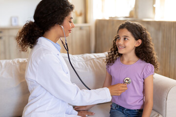 Friendly pediatrician woman checking lungs of little girl using stethoscope to examine breathing and heartbeat of young patient during home visit
