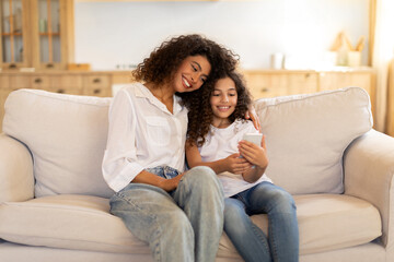 Happy mother and little daughter using smartphone sitting on sofa at home, scrolling social media,...
