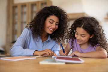 Caring young mom and her daughter sitting at table at home and making homework assignment together, tutor and girl child study learn do home task - obrazy, fototapety, plakaty